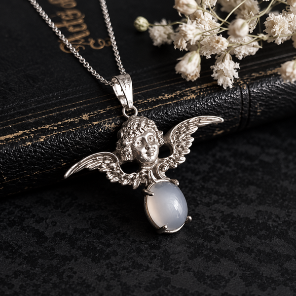 A silver angel pendant necklace with a detailed winged cherub head and an oval milky white stone, displayed on a dark textured surface next to dried white flowers and a black book.