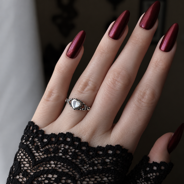A close-up of a hand with long, glossy dark red manicured nails wearing a silver ring with a heart-shaped stone, styled with a black lace glove.