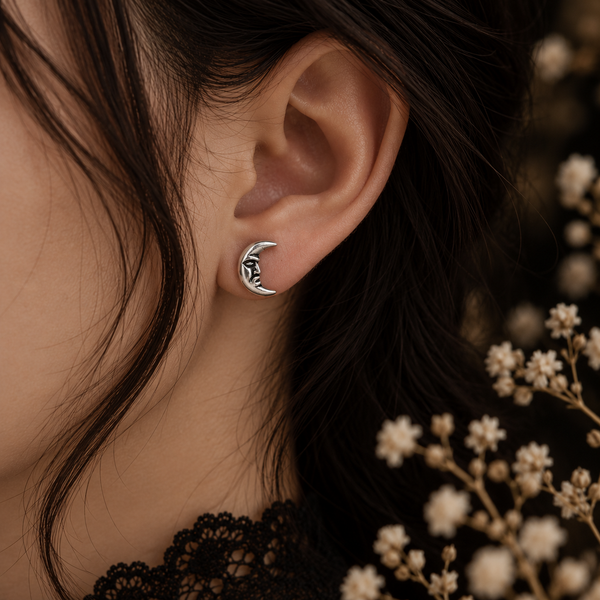 A close-up of a person's ear wearing a small crescent moon stud earring with a black leaf design inside, styled with dark hair and dried white flowers in the background.
