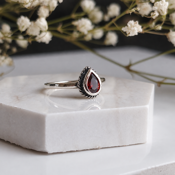 A silver ring with a teardrop-shaped red gemstone, displayed on a white marble block with white baby's breath flowers in the background.