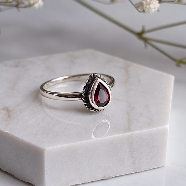 A silver ring with a teardrop-shaped dark red gemstone, displayed on a white marble hexagonal block with delicate baby's breath flowers in the background.