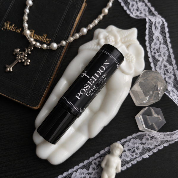A black perfume bottle roller labeled 'POSEIDON Citrus & Herbs' rests on a white sculptural hand-shaped dish, styled with a pearl necklace, black book, lace trim, clear crystal stones, and a small white figurine on a dark surface.