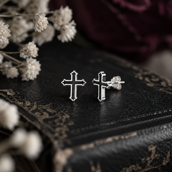 A pair of silver cross stud earrings displayed on a dark textured book, with dried white flowers in the foreground and a dark burgundy fabric background.