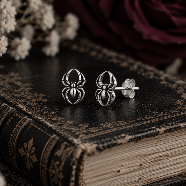 A pair of silver spider stud earrings displayed on an antique dark book, with dried white flowers and a dark burgundy rose in the soft-focus background.