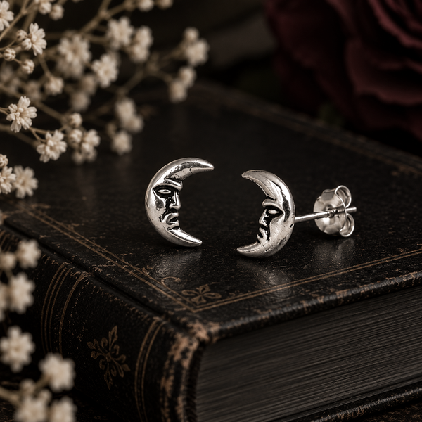 A pair of silver crescent moon stud earrings with engraved faces, displayed on a dark vintage book with dried white flowers in the foreground.