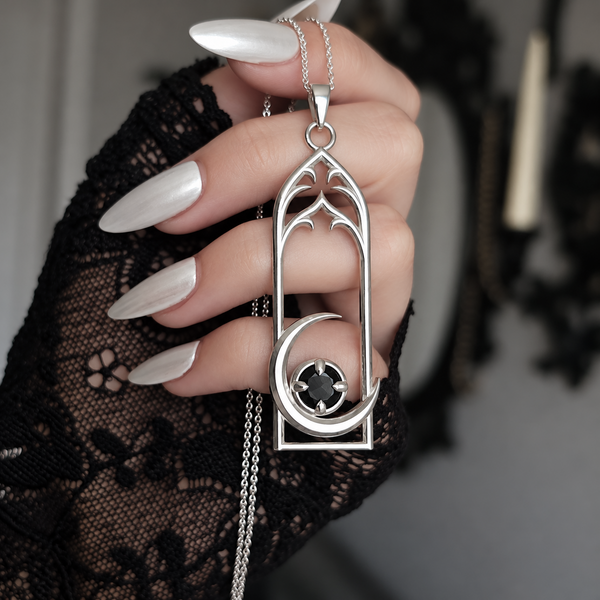 A hand with long white manicured nails and a black lace sleeve holds a silver gothic-style pendant necklace. The pendant features a pointed arch frame, a crescent moon, and a central black gemstone with four clear stones.