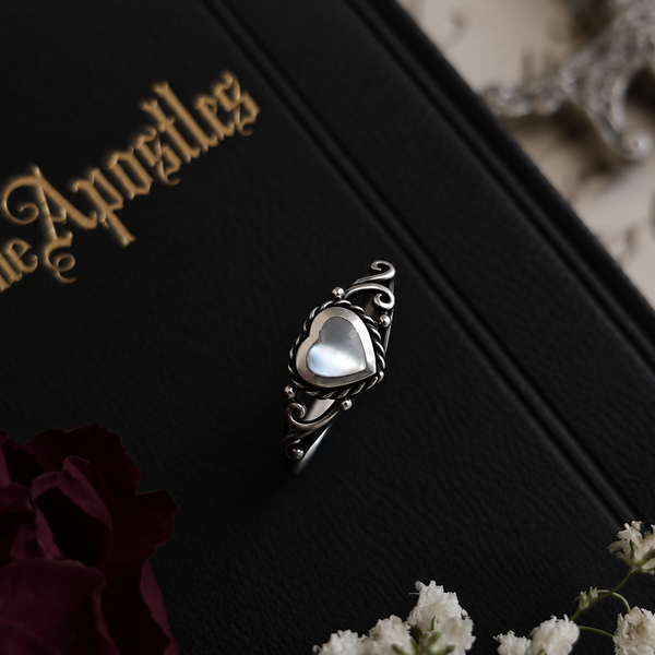 A silver ring with an ornate scrollwork band and a heart-shaped iridescent cabochon gemstone, displayed on a black book with gold lettering.
