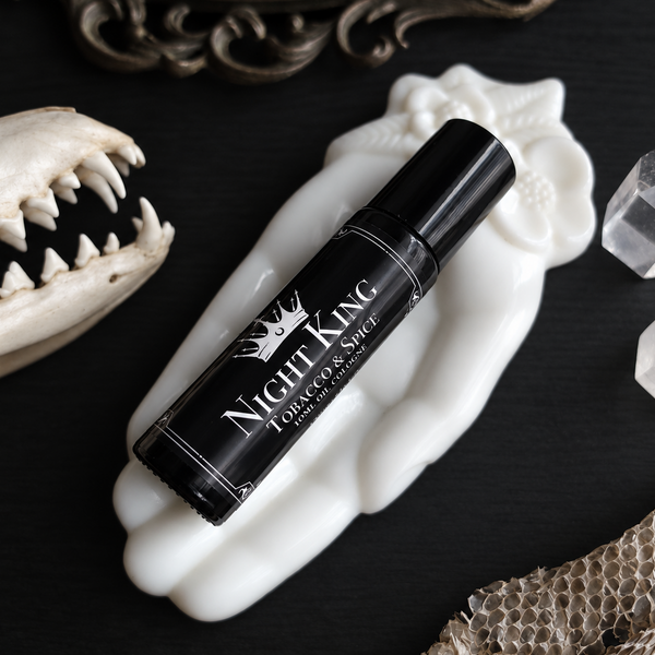 A black cylindrical rollerball bottle labeled 'Night King Tobacco & Spice' with white lettering, displayed diagonally on a white sculptural hand-shaped stand.