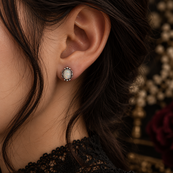 A close-up of a woman's ear wearing a small oval stud earring with a white pearl-like center and a silver-toned beaded border, against dark hair and a softly blurred dark background.