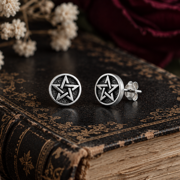 A pair of silver-toned circular stud earrings with a pentagram design, displayed on the edge of an antique book with dried flowers and a dark red rose in the background.