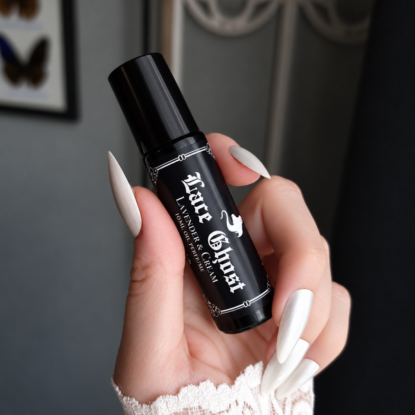 A hand with long white manicured nails holds a black roll-on bottle labeled 'Lace Ghost' against a dark gray background.