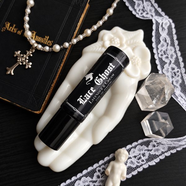 A black cylindrical perfume tube labeled 'Lace Ghost Lavender & Cream' rests on a white sculpted hand-shaped dish. The scene is styled with a pearl necklace, a black book, lace trim, clear crystal cubes, and a small white figurine on a dark surface.