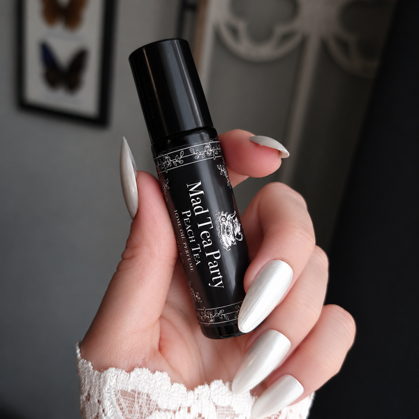 A hand with long white manicured nails holds a black perfume bottle labeled 'Mad Tea Party Peach Tea'  The background is a softly blurred indoor setting with a gray wall and framed butterfly artwork.