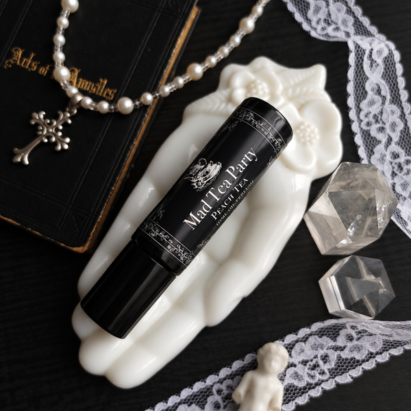 A black perfume bottle labeled 'Mad Tea Party Peach Tea' rests on a white sculpted hand-shaped dish. The scene is styled with a pearl necklace, a black book, lace trim, clear crystal stones, and a small white figurine on a dark background.