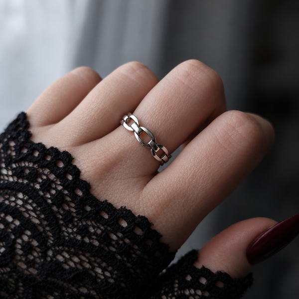 A close-up of a hand wearing a silver chain-link ring, with a black lace sleeve and dark red manicured nails visible.