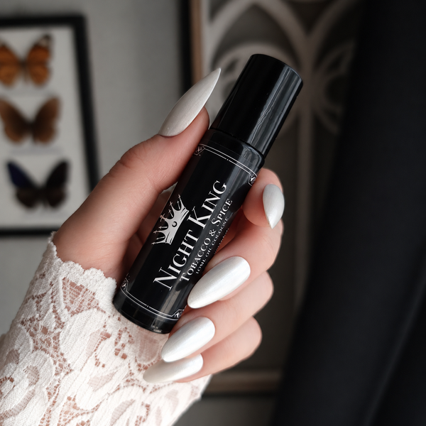 A hand with long white manicured nails holds a black cylindrical bottle labeled 'Night King' and 'Tobacco and Spice' against an indoor background with framed butterfly artwork.