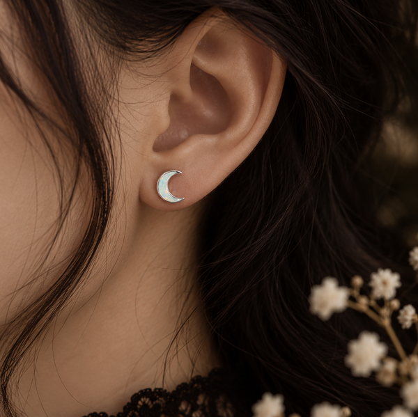 A close-up of a person's ear wearing a small crescent moon stud earring, with dark hair and delicate white flowers in the background.
