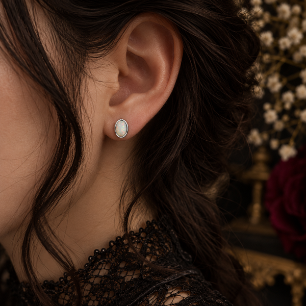 A close-up of a woman's ear wearing a small oval stud earring with a white iridescent shell-like center set in silver, against a dark background with floral decor.