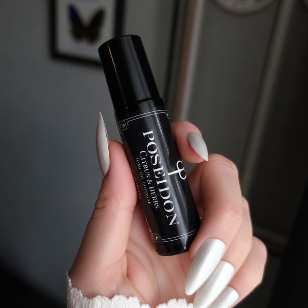 A hand with long, glossy white manicured nails holds a black perfume roller bottle labeled 'POSEIDON Citrus & Herbs'. The background is a softly blurred indoor setting with gray walls and framed wall art.