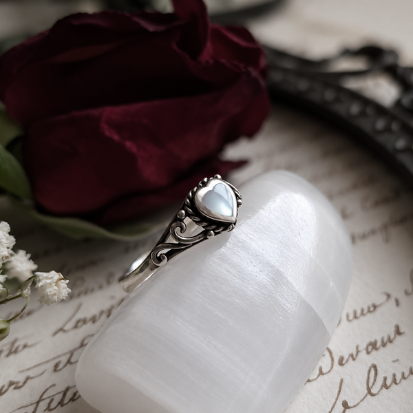 An ornate silver ring with a heart-shaped white mother-of-pearl gemstone, displayed on a white stone holder with a dark red rose and vintage script paper in the background.