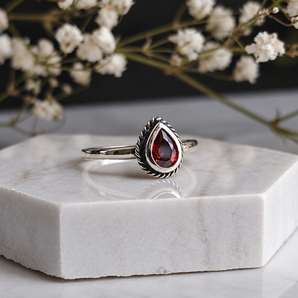 A silver ring with a teardrop-shaped dark red gemstone, displayed on a white marble hexagonal block with white baby's breath flowers in the background.