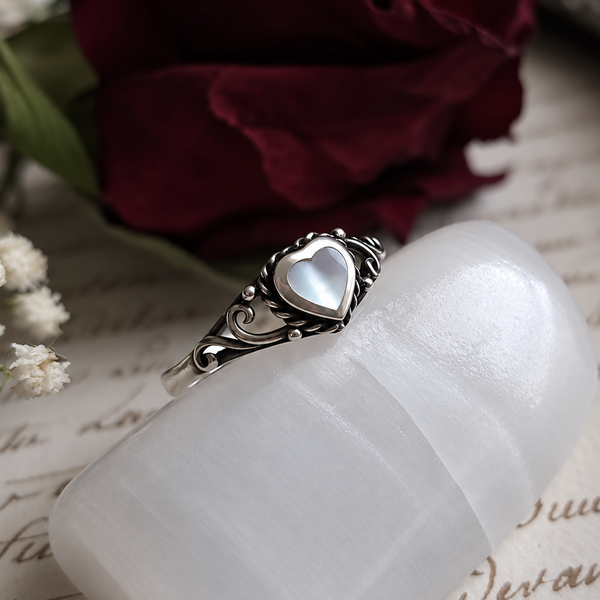A silver ring with a mother-of-pearl heart-shaped center stone, displayed on a white selenite holder with a dark red rose and vintage script paper in the background.