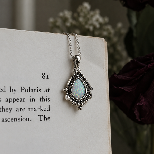 A silver-toned teardrop pendant necklace with an iridescent white opal center, draped over the corner of an open book with blurred flowers in the background.