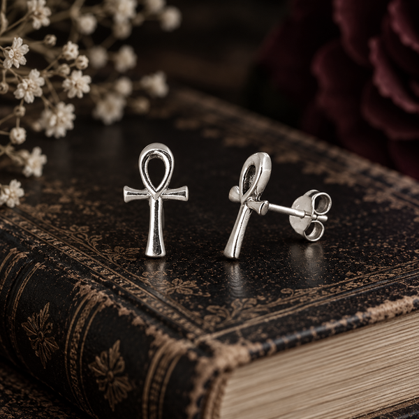 A pair of silver ankh symbol stud earrings displayed on an antique dark brown book, with dried white flowers in the background.